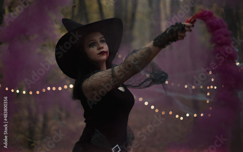 Portrait in the woods with a sexy woman with a hat on her head and a black dress, made up with fake blood. She holds a colorful fumigant in her hand.