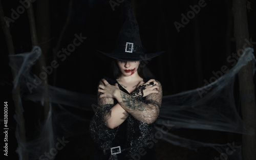 Sexy halloween portrait of brunette haired woman with hat on her head, makeup with fake blood. Dark forest background.
