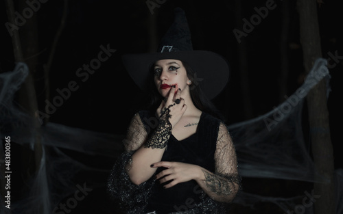 Sexy halloween portrait of brunette haired woman with hat on her head, makeup with fake blood. Dark forest background.