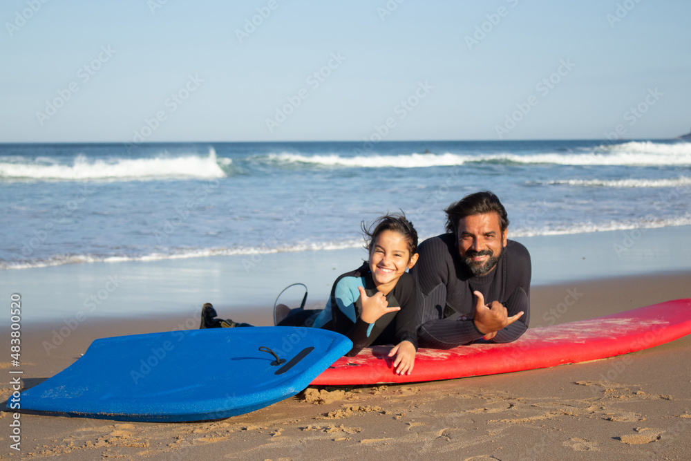 Obraz premium Caring father with mechanical leg with daughter on beach. Mid adult man and little dark-haired girl lying on surfboards, showing shaka signs with fingers. Family, leisure, active lifestyle concept