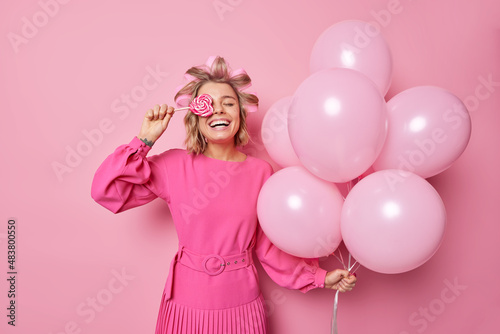 Happy woman has festive mood smiles broadly holds sweet candy over eye wears dress applies hair rollers poses with helium balloons enjoys party time isolated on pink background. Celebration.
