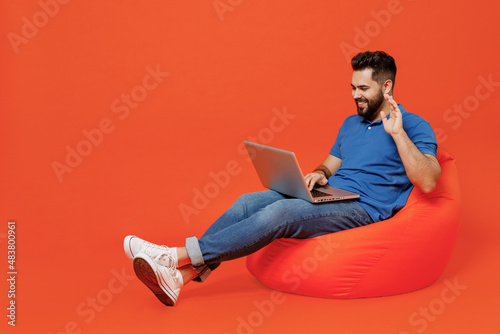 Full body young smiling friendly happy fun man wear basic blue t-shirt sit in bag chair hold use work point on laptop pc computer talk by video call waving hand isolated on plain orange background.