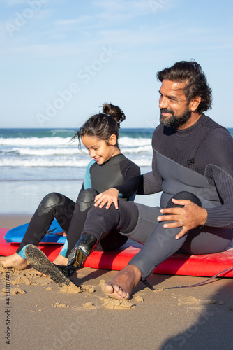 Bright father with mechanical leg with daughter on beach. Mid adult man and little dark-haired girl sitting on surfboards, resting after training. Family, leisure, active lifestyle concept