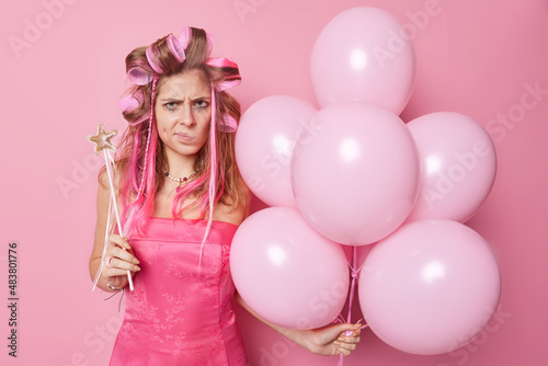 Displeased woman purses lips looks angrily doesnt like something comes on party feels frustrated poses with inflated balloons and magic wand isolated over pink background. Negative emotions.