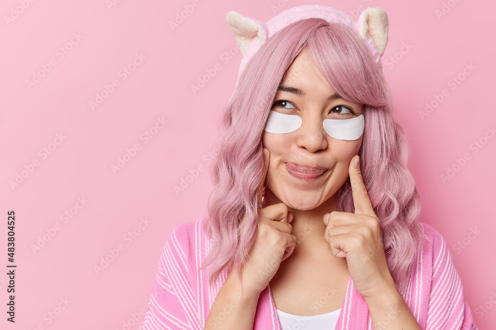 Pleased young Asian woman keeps index fingers on cheeks licks lips with tongue applies beauty patches under eyes to remove wrinkles wears headband isolated over pink background blank copy space