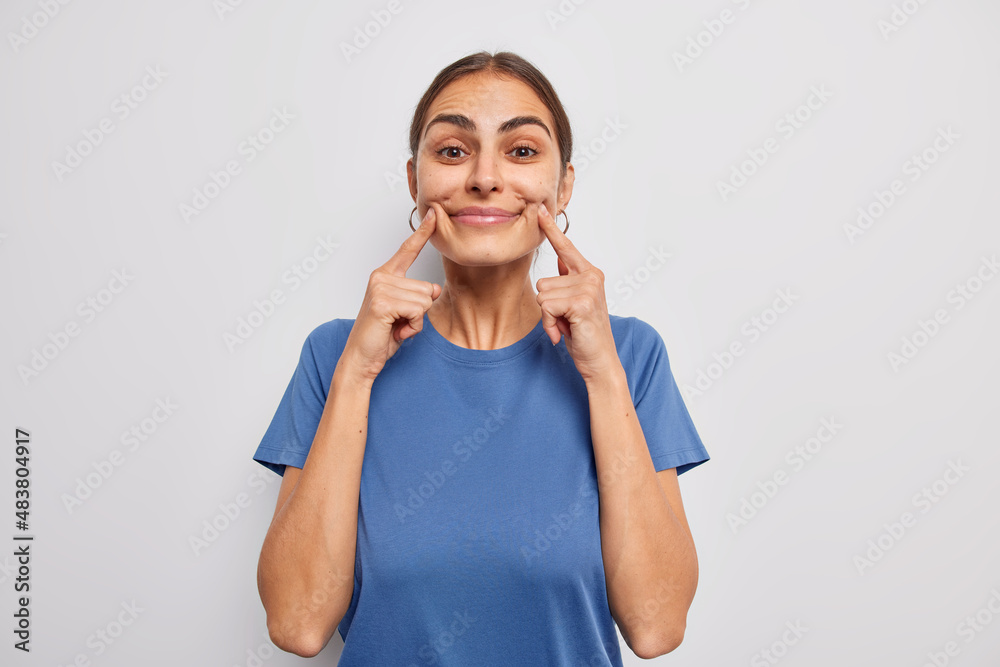 Beautiful dark haired woman keeps fingers near corner of lips tries to have happy expression forces smile dressed in casual blue t shirt isolated over white background. Look at my cheerful face Beautiful dark haired woman keeps fingers near corner of lips tries to have happy expression forces smile dressed in casual blue t shirt isolated over white background. Look at my cheerful face