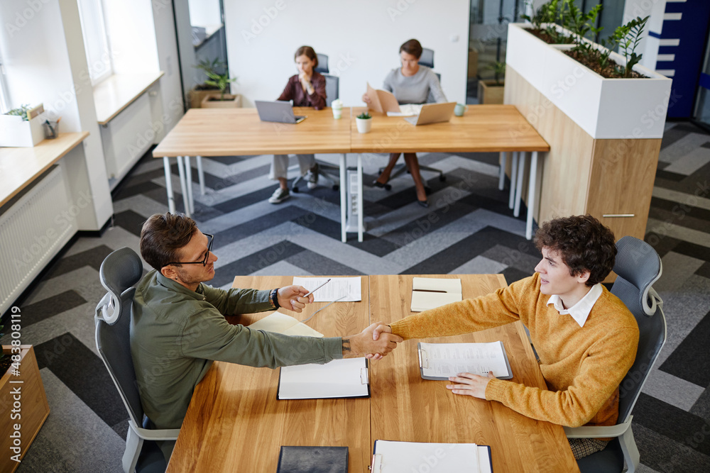 High angle portrait of HR manager shaking hands with young man at job interview in office, copy space High angle portrait of HR manager shaking hands with young man at job interview in office, copy space