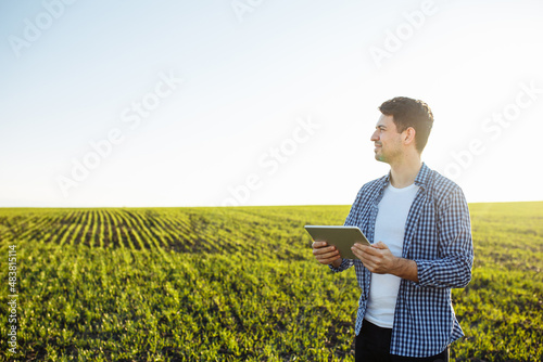 Agronomist stands in the young green wheat field with the tablet in his hands and sends groth progress data to the server for future analysis. Spring seeding concept.