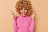Happy joyful woman reacts on good news keeps palms raised and laughs positively wears casual pink turtleneck gestures actively poses against brown background. Positive human reactions concept