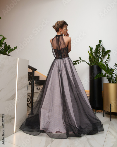 Elegant woman with hairstyle wearing white floor length evening gown with black chiffon decor stands near marble stairs in spacious hallway photo