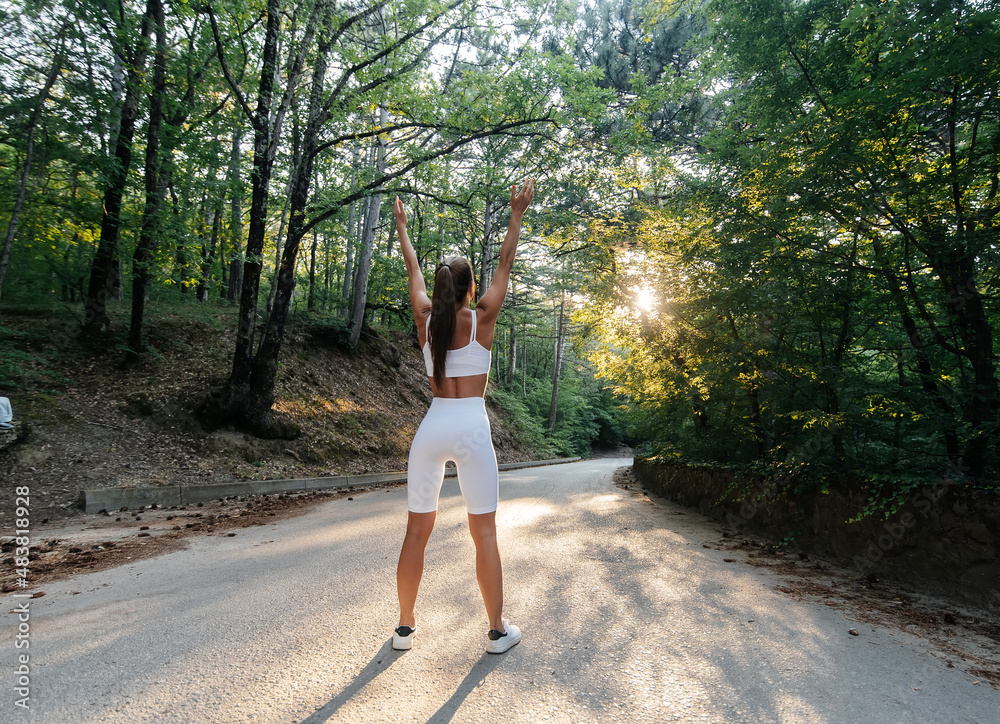 A young beautiful girl poses with her back before running training, on the road in a dense forest, during sunset. A healthy lifestyle and running in the fresh air. A young beautiful girl poses with her back before running training, on the road in a dense forest, during sunset. A healthy lifestyle and running in the fresh air.