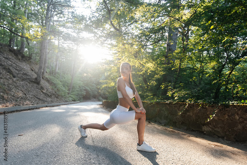 A young beautiful girl makes lunges and warm-up before running training, on the road in a dense forest, during sunset. Healthy lifestyle and running in the fresh air. Outdoor training.