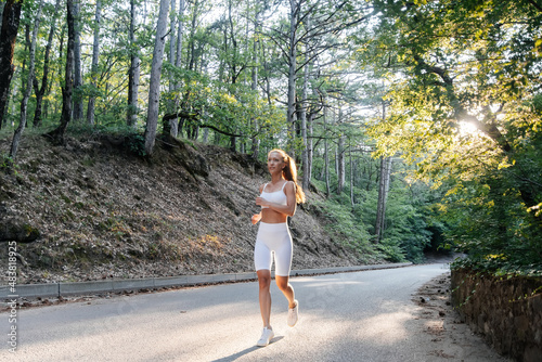 A young beautiful girl in white sports clothes runs along the road in a dense forest during sunset. I do sports in the fresh air. Healthy lifestyle. Outdoor training.