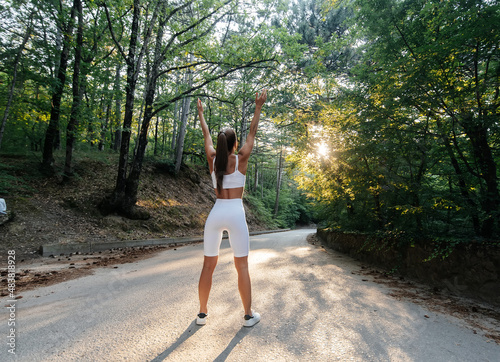 A young beautiful girl poses with her back before running training, on the road in a dense forest, during sunset. A healthy lifestyle and running in the fresh air.