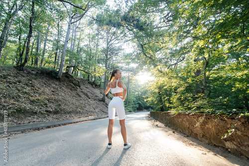 A young beautiful girl poses with her back before running training, on the road in a dense forest, during sunset. A healthy lifestyle and running in the fresh air.