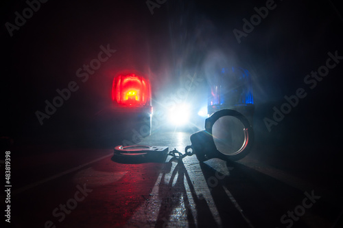 Police raid at night and you are under arrest concept. Silhouette of handcuffs with police car on backside. Image with the flashing red and blue police lights at foggy background.