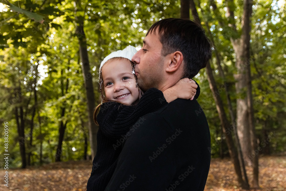 Dad and daughter are hugging and the daughter is looking at the camera. Summer park. Green trees. The concept of a young family. The decoration with stars is woven into the girl's hair. Dad and daughter are hugging and the daughter is looking at the camera. Summer park. Green trees. The concept of a young family. The decoration with stars is woven into the girl's hair.