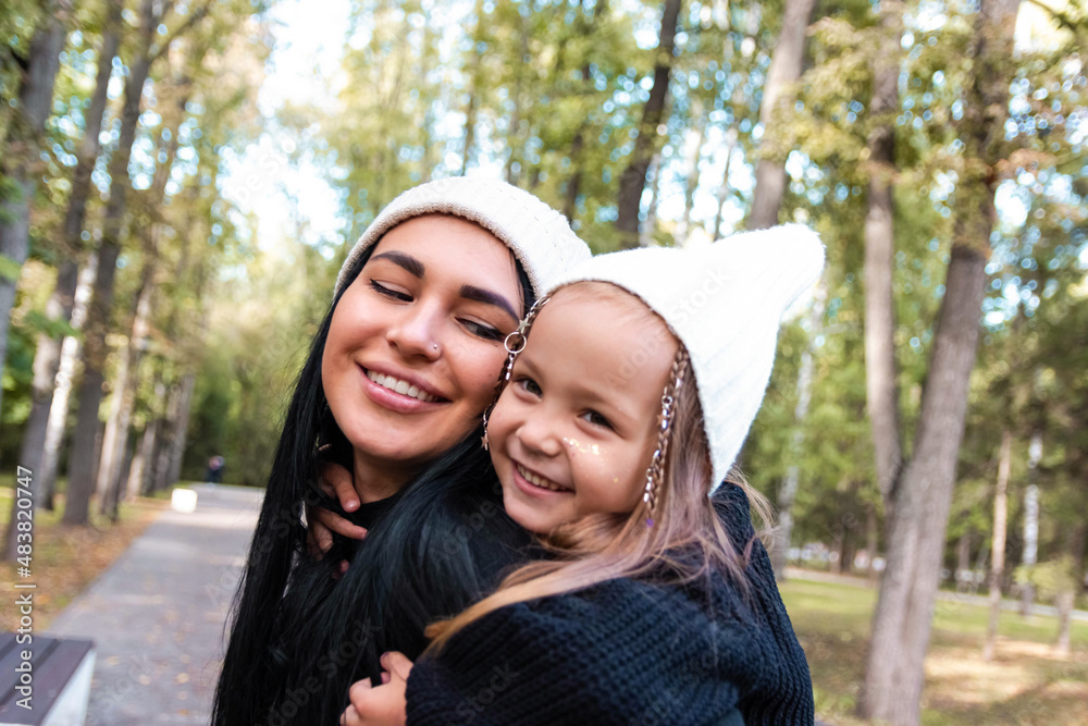 Fototapeta premium Mom and daughter. The little daughter sits on her back. Tenderness of mother and daughter. Summer park. Green trees. The concept of a young family. Braided stars on the hair.