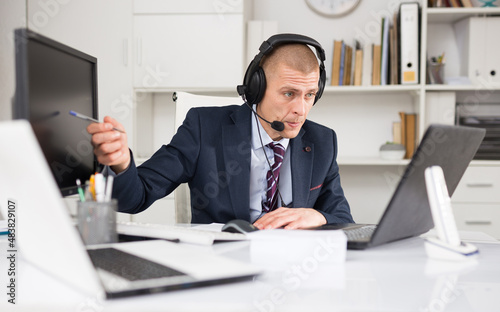 Positive businessman with headphones and microphone having video call on laptop in modern office