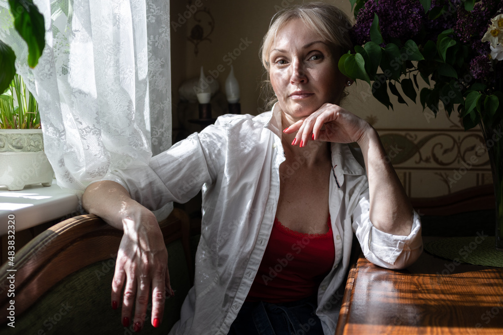 Blond middle aged woman in white shirt is sitting near the window