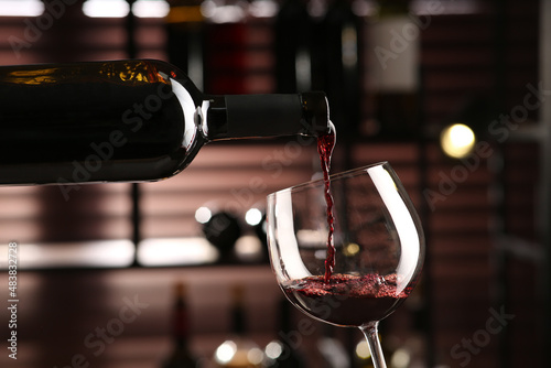 Pouring red wine from bottle into glass on blurred background, closeup photo