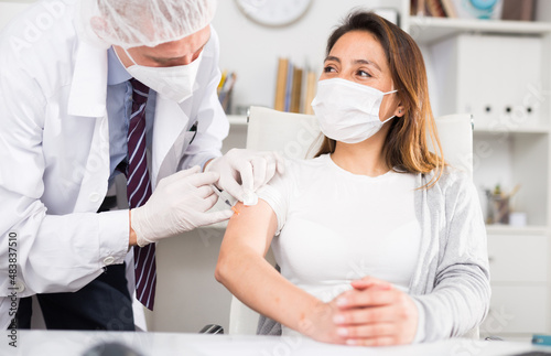 Doctor in white coat and protective mask giving vaccine injection to woman manager in office