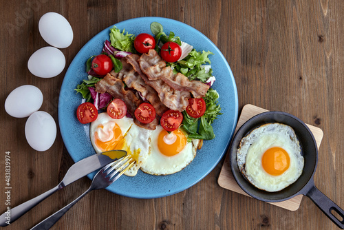 Fried eggs with bacon, lettuce and cherry tomatoes on a dark wooden plate