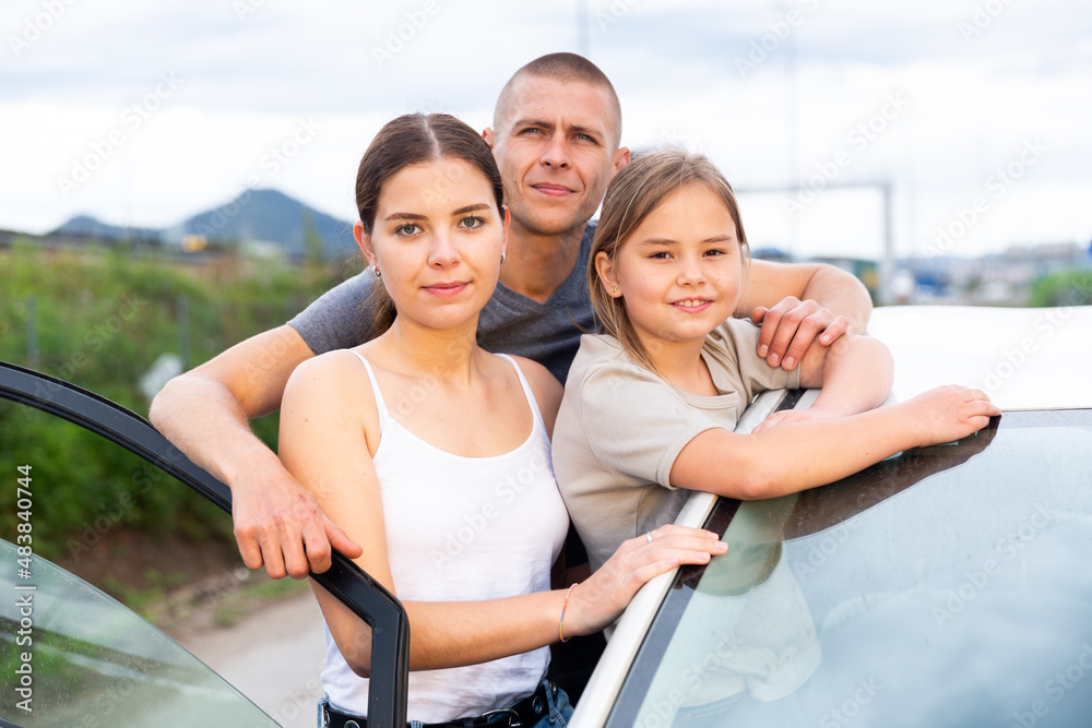 Friendly young family of three posing outside next to car