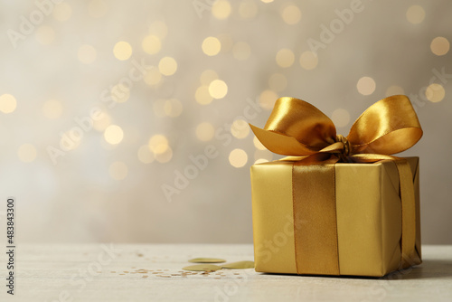 Beautiful golden gift box on light table against blurred festive lights, space for text