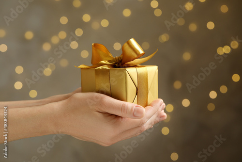 Woman holding beautifully wrapped gift box against blurred festive lights, closeup photo