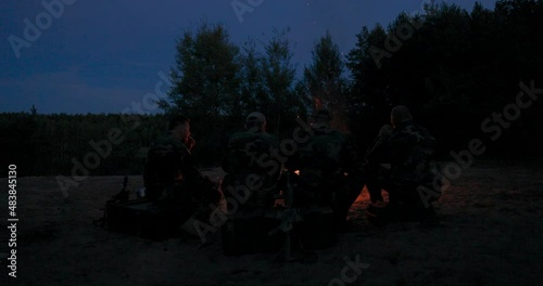 A team of men in camouflage clothing is sitting on a hill in the field at night, warming themselves by a lit campfire, frying sausages for dinner, a manly outing, relaxing after work, service, hunting photo