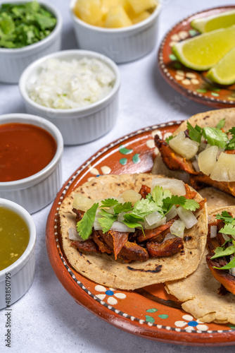 Traditional Mexican street food called "Tacos al pastor" prepared with pork, tortillas, pineapple, cilantro, onion, green sauce, red sauce and lemons. On white texture background.