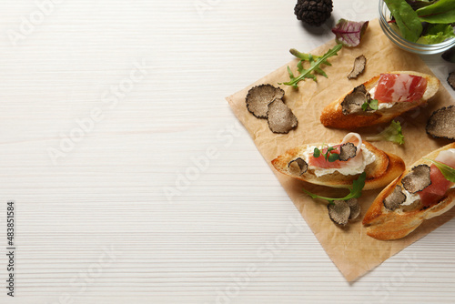 Delicious bruschettas with cheese, prosciutto and slices of black truffle on white wooden table, flat lay. Space for text