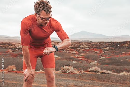 Tired runner man checking wearable technology sports smartwatch during outdoor training. Sport athlete looking at watch during workout on desert trail. Man wearing sunglasses, compression outfit