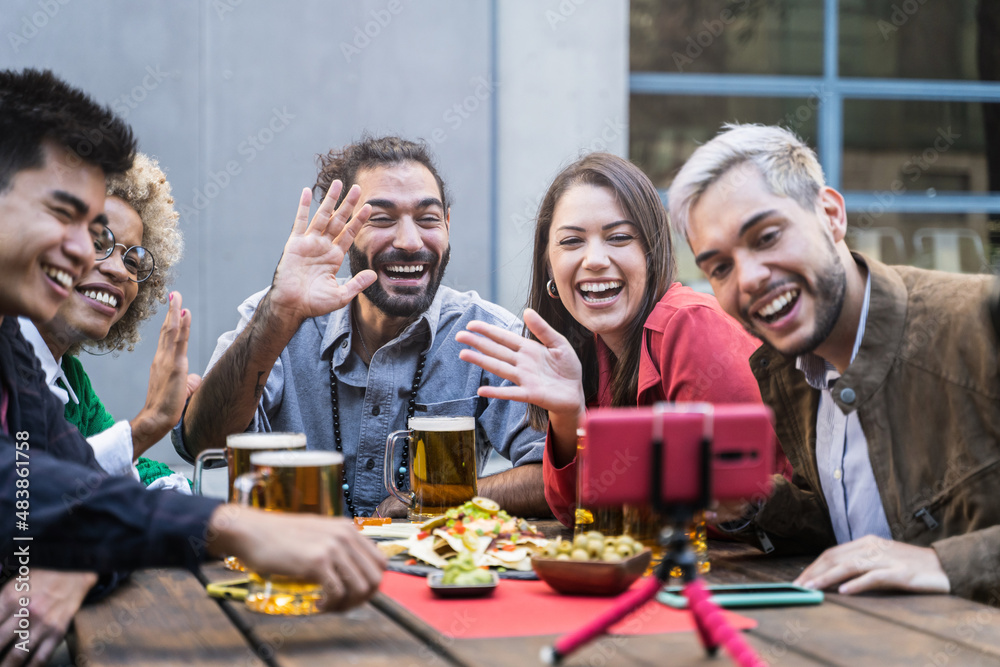 Obraz premium Group of young friends make Video Call in outdoors bar. Beautiful Young Diversity People drinking beer