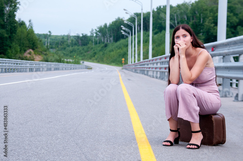 A girl is sitting on a suitcase on the side of an empty road waiting for a car