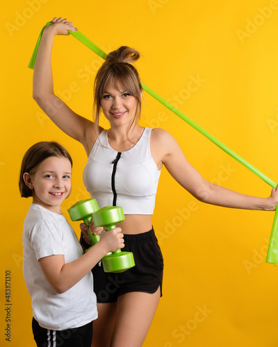 Healthy fit mom and daughter wearing sport wear in studio using resistive exercise band and dumbbells against yellow background. Active lifestyle. Sporty Family together online workouts at home. photo