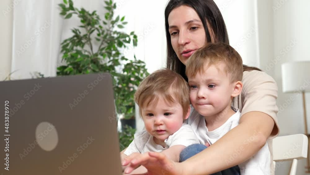 Happy and tired mother working from home, working at the table with a laptop, two sons are sitting in her arms. The concept of motherhood and parenting. Happy and tired mother working from home, working at the table with a laptop, two sons are sitting in her arms. The concept of motherhood and parenting.
