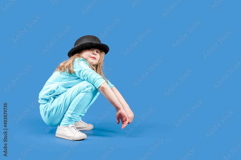 Cheerful teenage schoolboy grimaces into the camera. Fidget. A lovely girl in a black hat and tracksuit sits on a blue background. Copy space. Cheerful teenage schoolboy grimaces into the camera. Fidget. A lovely girl in a black hat and tracksuit sits on a blue background. Copy space.
