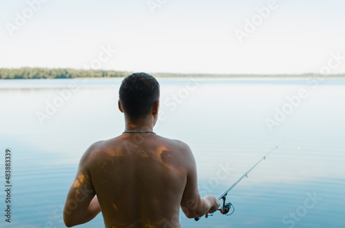 Male fisherman fishing on sunny summer day outdoors, waist length, back view. Caucasian man holding spinning rod standing on shore of lake in nature. Sport and hobby, leisure and recreation concept
