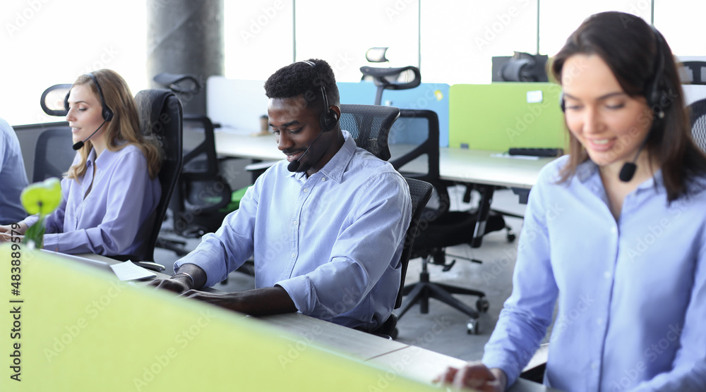 Smiling african male call-center operator with headphones sitting at modern office with collegues on the backgroung, consulting online. Smiling african male call-center operator with headphones sitting at modern office with collegues on the backgroung, consulting online.