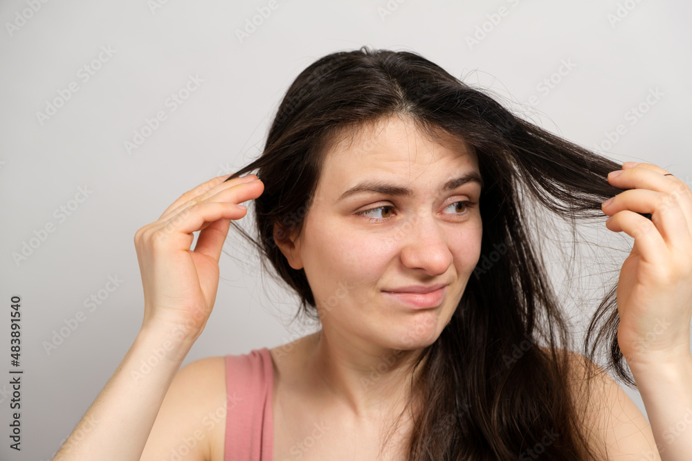 A beautiful brunette woman without makeup with dirty oily hair holds her hands on her head with a dissatisfied look A beautiful brunette woman without makeup with dirty oily hair holds her hands on her head with a dissatisfied look