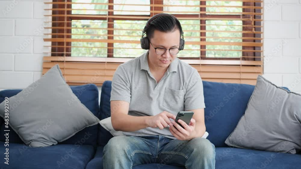 Young man wearing headphones and holding smartphone in comfortable living room. Young male listening music audio sound and happy expression at home. Young man wearing headphones and holding smartphone in comfortable living room. Young male listening music audio sound and happy expression at home.