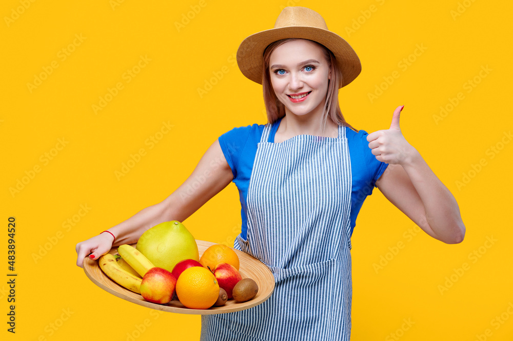 Smiling female gardener holding fruits banana apple orange kiwi grapefruit in wooden plate dressed in straw hat and an apron and show thumb up gesture on yellow background Smiling female gardener holding fruits banana apple orange kiwi grapefruit in wooden plate dressed in straw hat and an apron and show thumb up gesture on yellow background