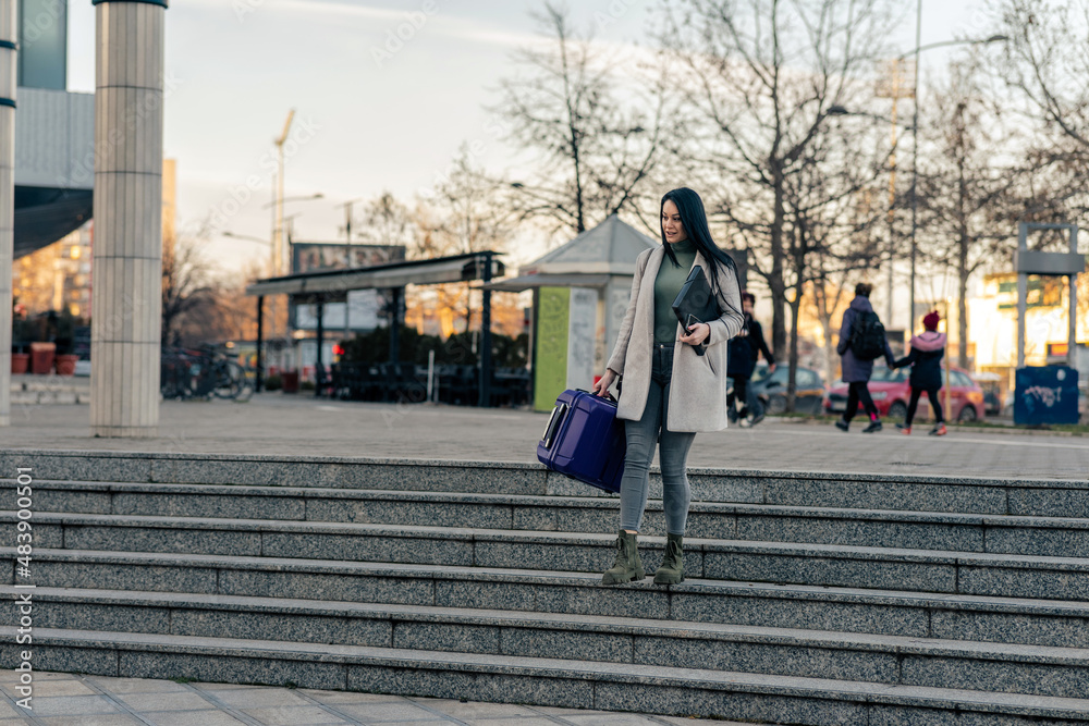 Full length portrait of a smiling successful business woman carrying a suitcase, documents and a mobile phone.