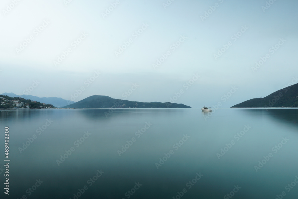 Small ship in the fog on the surface of water with mountains on the background. Bleu and gray tones