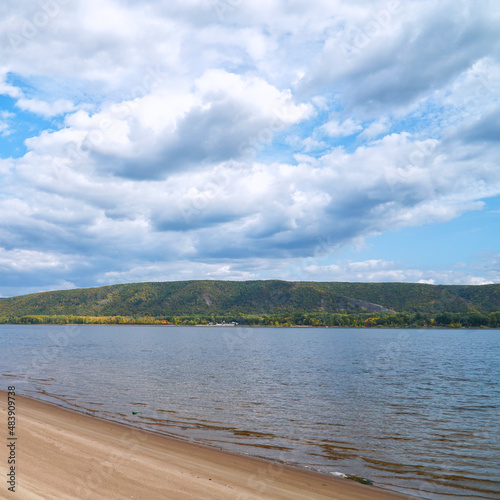 The sandy shore of the big Volga river against the background of blue cloudy sky. On the opposite bank of the Zhiguli Mountains