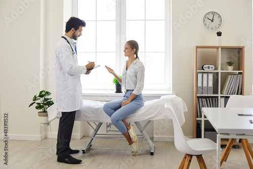 Patient talking to a doctor at a modern clinic or hospital. Male physician in a white coat giving a consultation to a young woman who is sitting on the medical couch in the exam room