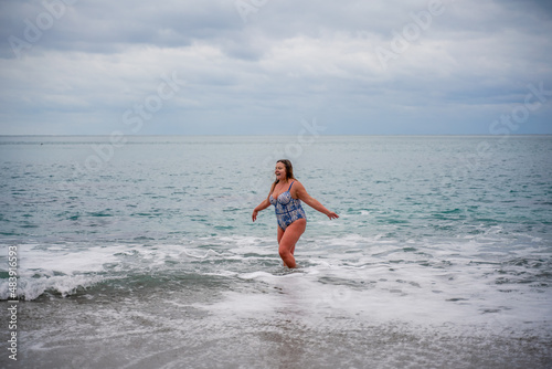 A fat woman in a bathing suit enters the water during the surf. Alone on the beach, Gray sky in the clouds, swimming in winter.