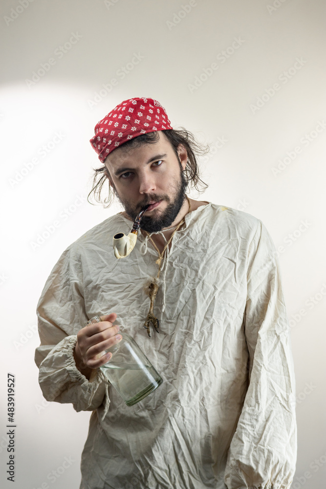 Portrait of a young dark-haired man with a beard, wearing a red cloth bandana on his head and an old shirt with a bottle of rum and a smoking pipe in his teeth on a light background. Pirate Costume, Portrait of a young dark-haired man with a beard, wearing a red cloth bandana on his head and an old shirt with a bottle of rum and a smoking pipe in his teeth on a light background. Pirate Costume,